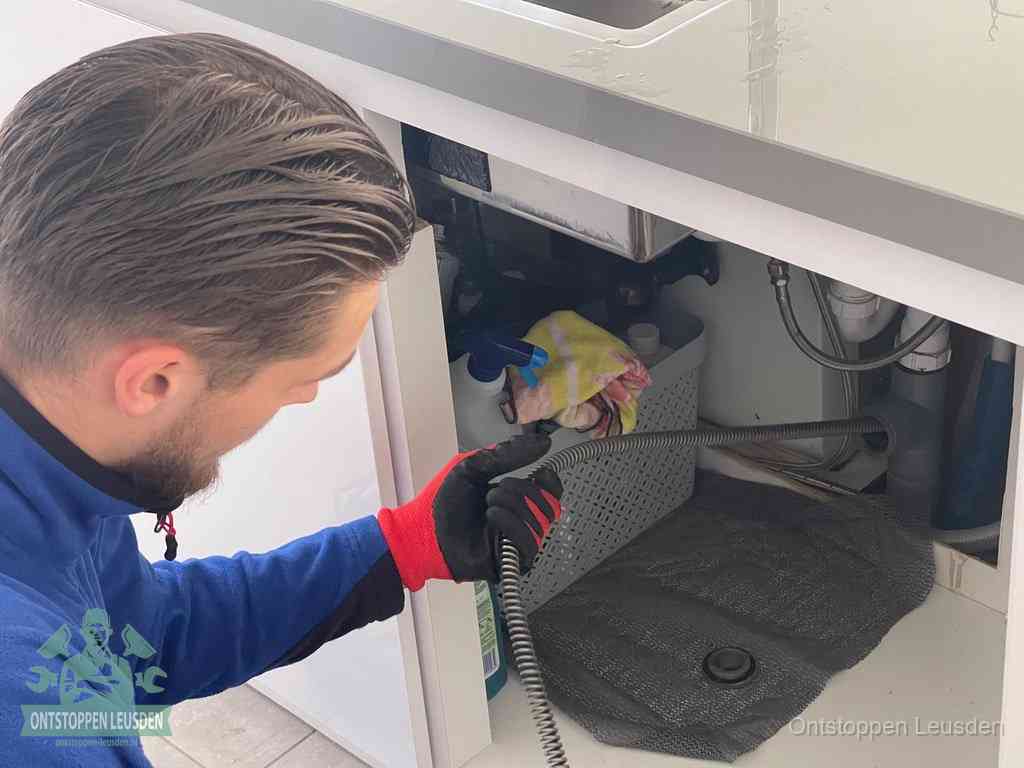 Een monteur voert een flexibele ontstoppingsveer in onder een keukenkastje om een verstopping voorbij de sifon te bereiken in Leusden
