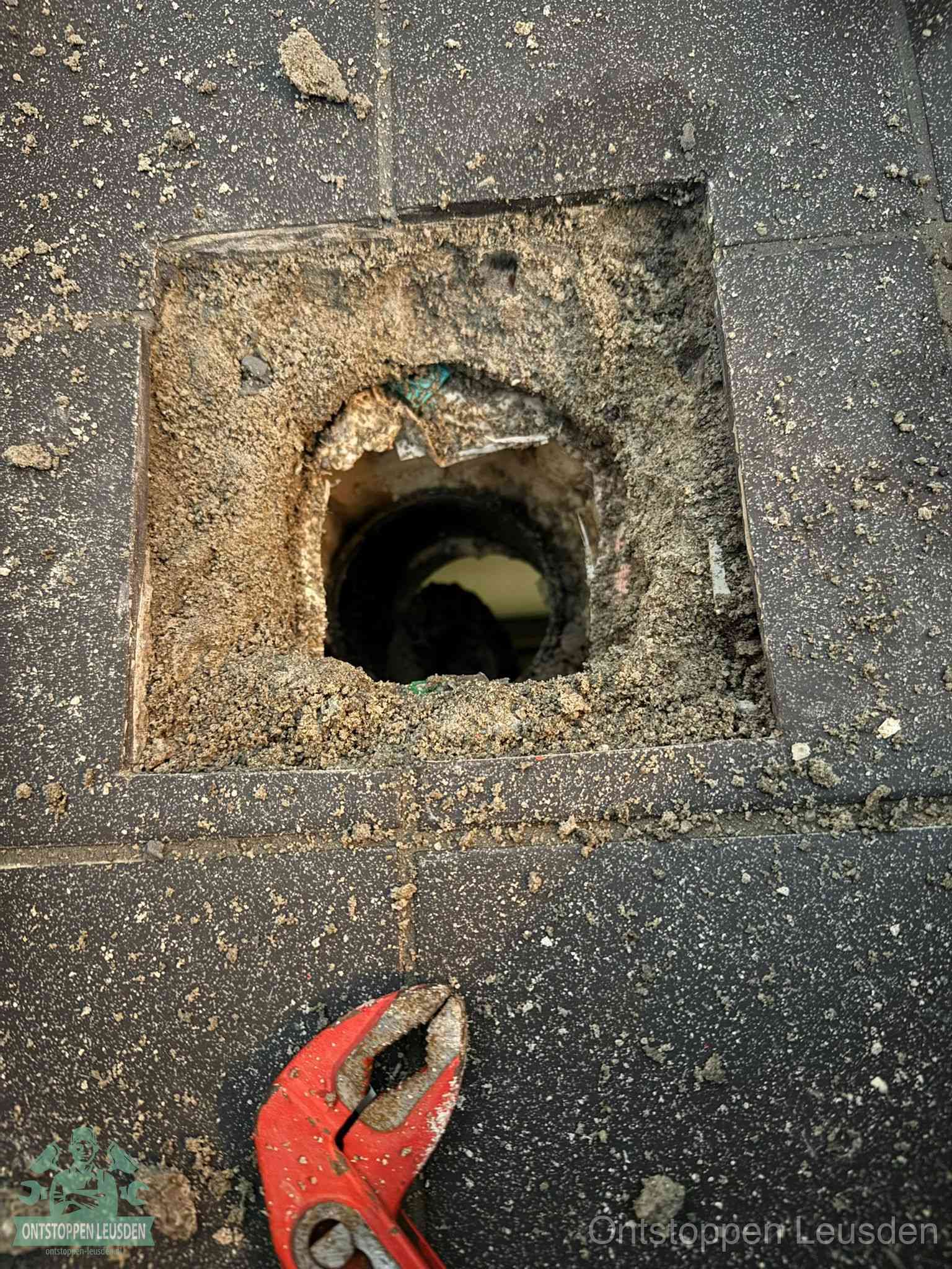 Een geopend rioolputje in een betontegelvloer met losgemaakt rooster en een zichtbare ronde afvoerbuis, het achterhalen waar de afvoerverstopping zit in Leusden