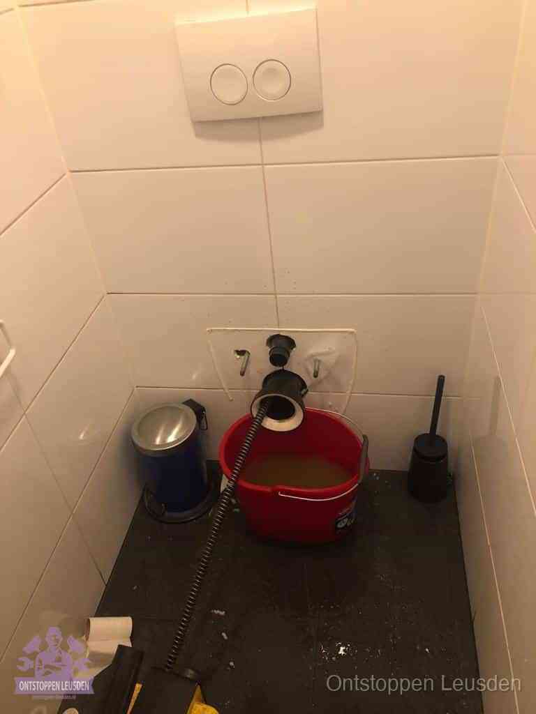 Een ontstopte wand-wc met verwijderde toiletpot waar een ontstoppingsveer in de wanduitgang verdwijnt, het achterhalen waar de verstopping zit in Leusden