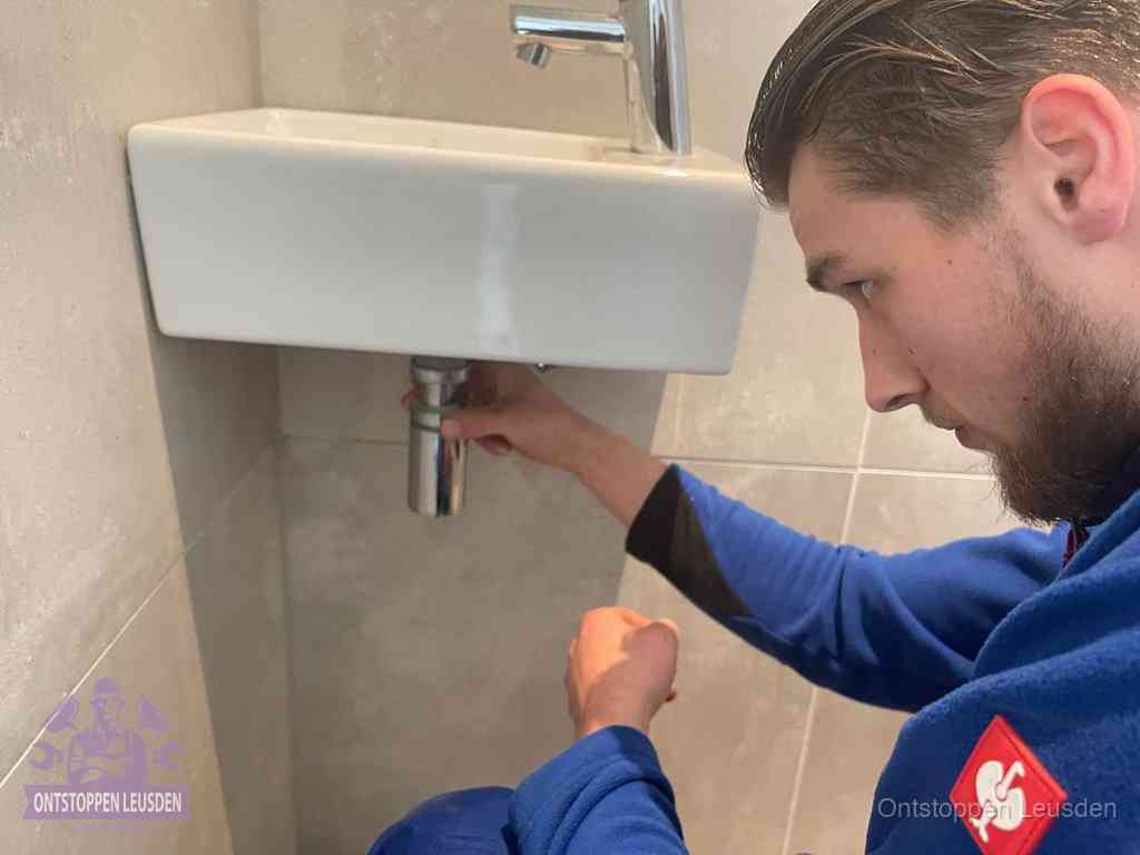 Een monteur draait met de hand de zwanenhalssifon onder een fonteinwasbak in de toiletruimte los om de bron van rioollucht in Leusden te vinden