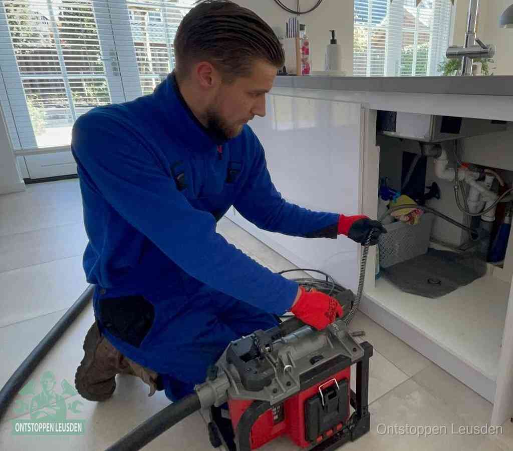 Een monteur met rode handschoenen knielt in een keuken en voert een elektrische ontstoppingsveer in tijdens het professioneel afvoer ontstoppen in Leusden